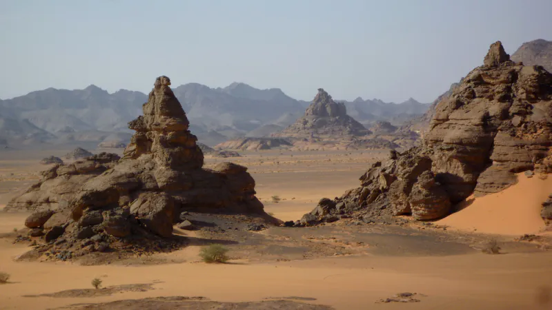 Weathered brown rock towers on a desert plain: a tall rock tower on dark boulders at the left, a rocky ridge with an orange dune at the right, flat sand between them and grey mountain ranges in the haze behind.