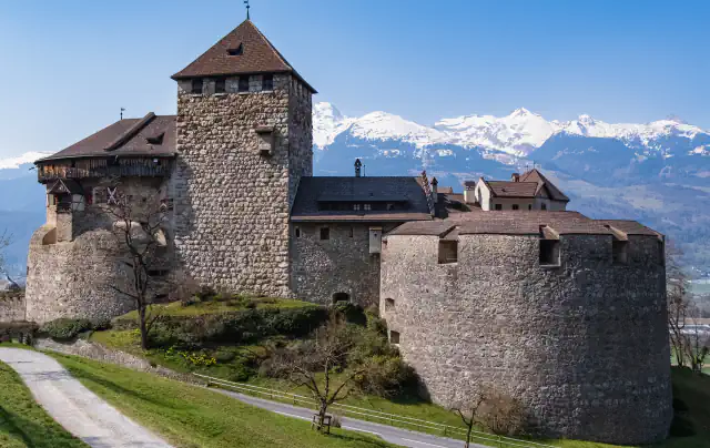 A stone castle with a tall square tower, a lower wing with a wooden upper storey and a broad round bastion stands above a road, with snow-covered mountains behind it under a clear blue sky