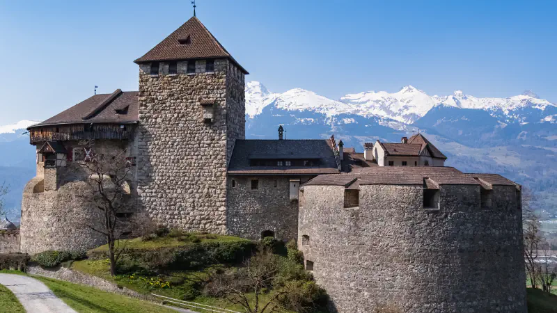 A stone castle with a tall square tower, a lower wing with a wooden upper storey and a broad round bastion stands above a road, with snow-covered mountains behind it under a clear blue sky