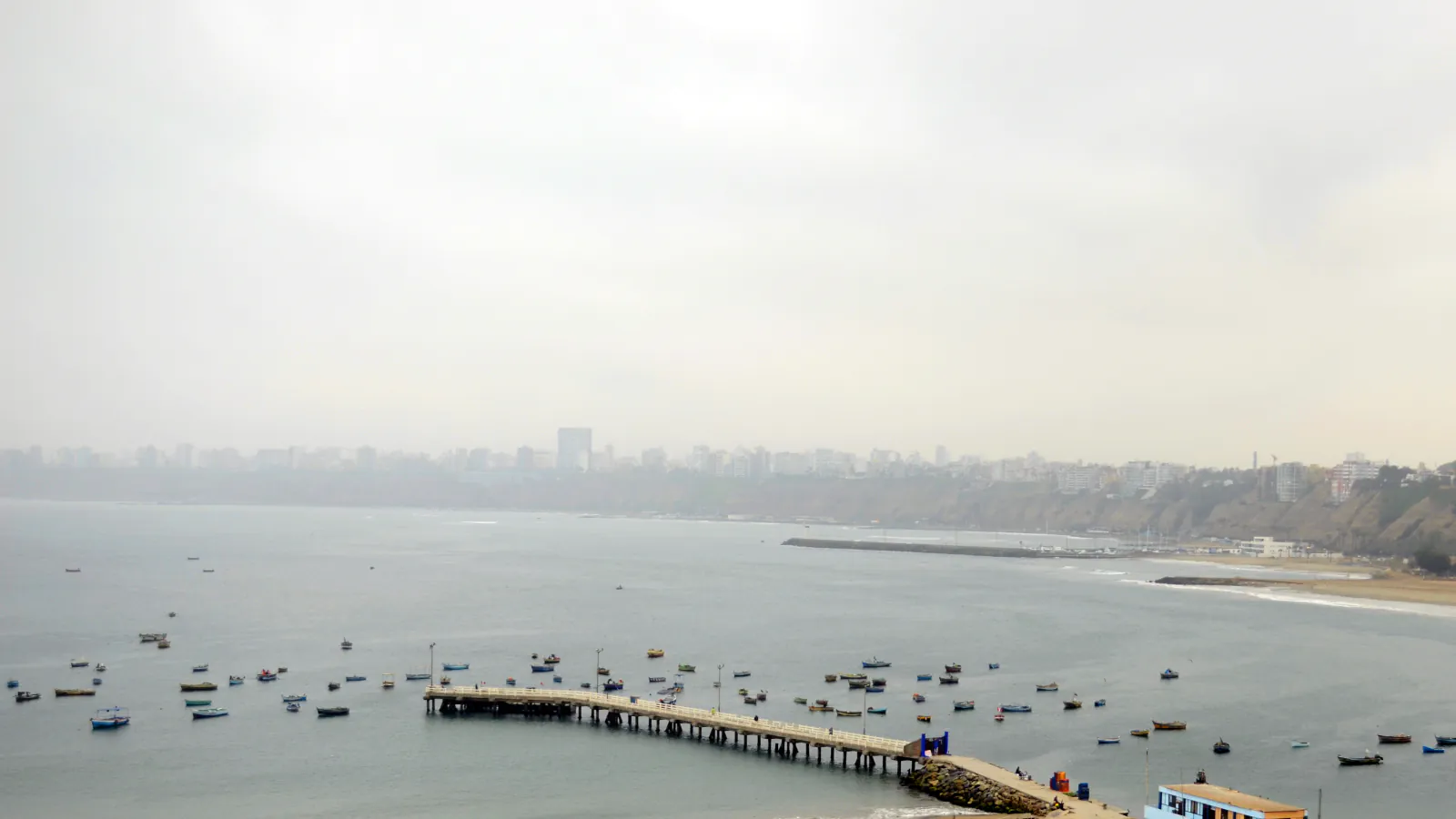 Small boats and a pier in a bay, with Lima’s hazy coastline and skyline in the distance