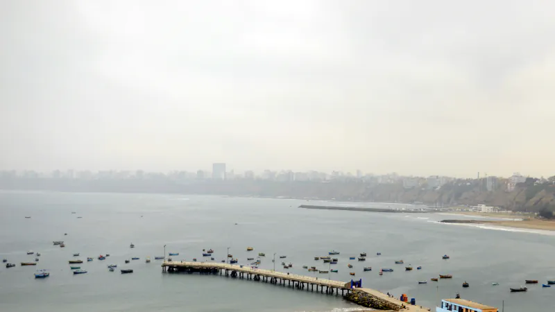 Small boats and a pier in a bay, with Lima’s hazy coastline and skyline in the distance