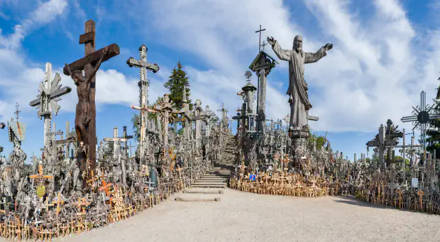 The Hill of Crosses near Šiauliai: many wooden and metal crosses on both sides of a gravel path and wooden steps climbing the hill, a large crucifix at left and a tall carved figure of Christ with open arms at right, under a blue sky with thin cloud