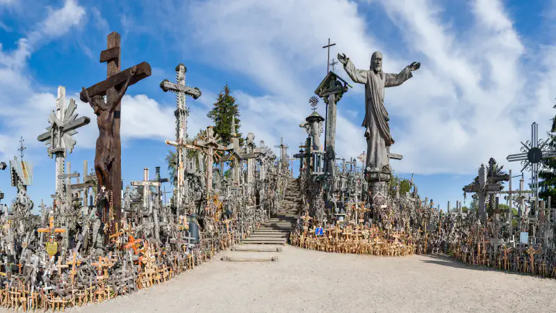 The Hill of Crosses near Šiauliai: many wooden and metal crosses on both sides of a gravel path and wooden steps climbing the hill, a large crucifix at left and a tall carved figure of Christ with open arms at right, under a blue sky with thin cloud