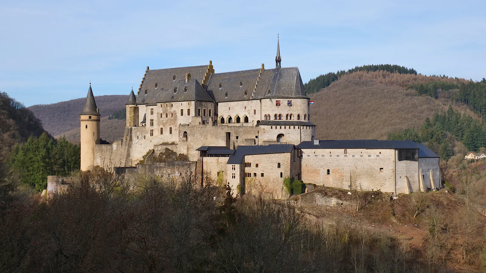 A large stone castle with slate roofs and a round tower at its left end stands on a rocky hilltop in front of wooded hills, under a pale blue winter sky