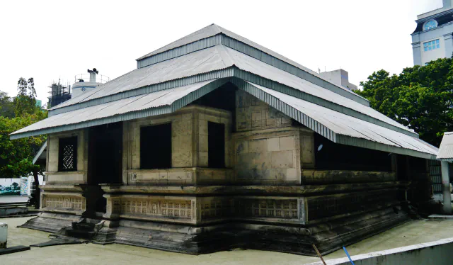 A low coral-stone mosque with carved panels along its base and a tiered roof of corrugated metal, seen from a corner, with trees and buildings behind