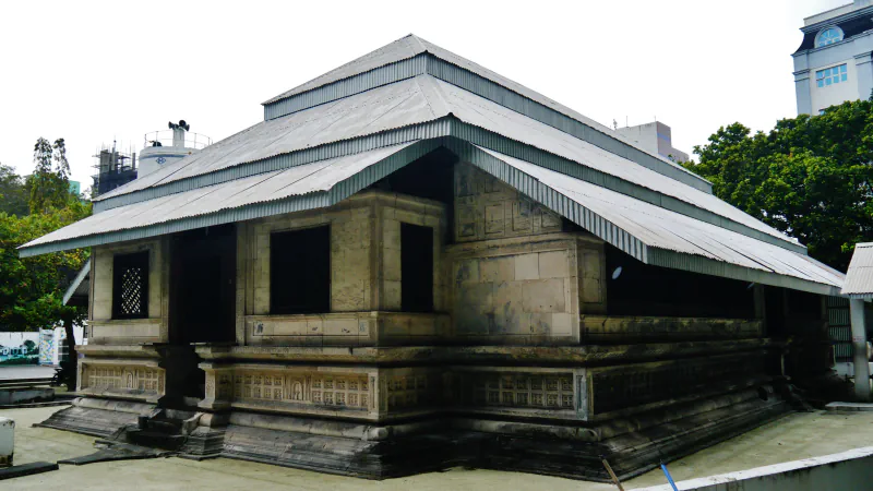 A low coral-stone mosque with carved panels along its base and a tiered roof of corrugated metal, seen from a corner, with trees and buildings behind