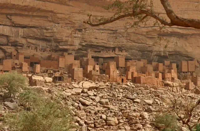 Flat-roofed earth buildings standing in rows below a tall banded sandstone cliff, among boulders and small trees, with a bare branch across the top right