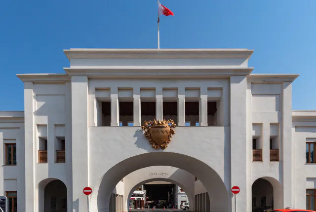 A white gate building with a broad central arch, a golden emblem and a flag on the roof under a clear sky.