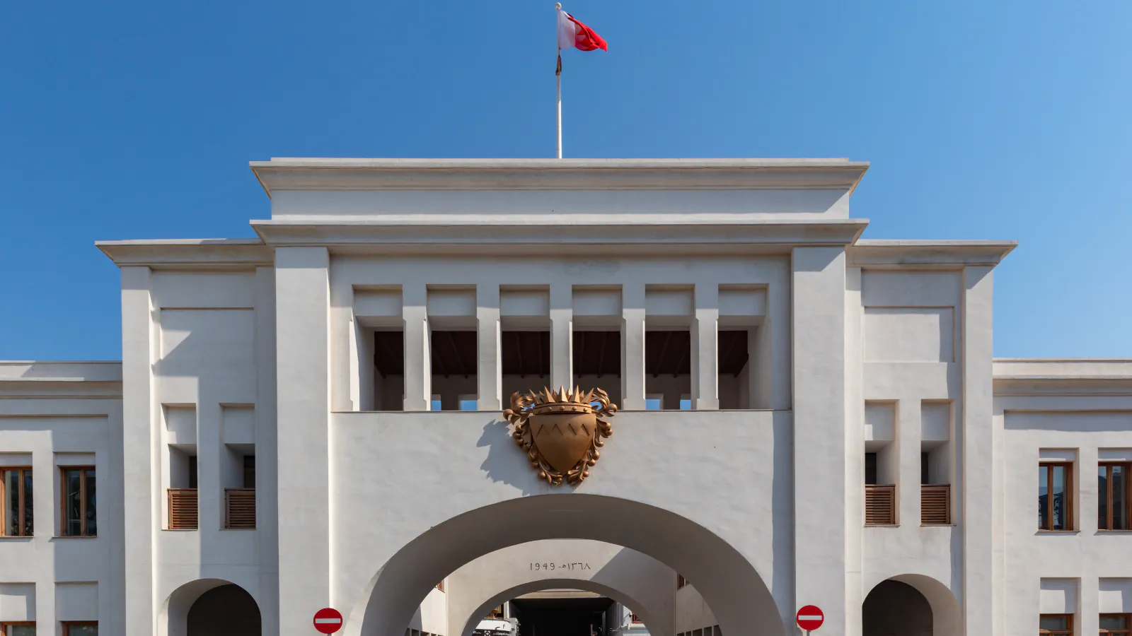 A white gate building with a broad central arch, a golden emblem and a flag on the roof under a clear sky.