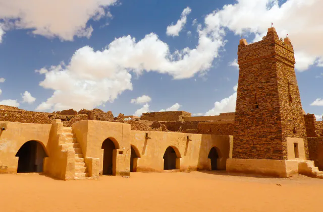 The square stone minaret of the mosque in Chinguetti rising beside low sand-coloured walls with arched doorways, with a sandy courtyard in the foreground and white clouds in a blue sky