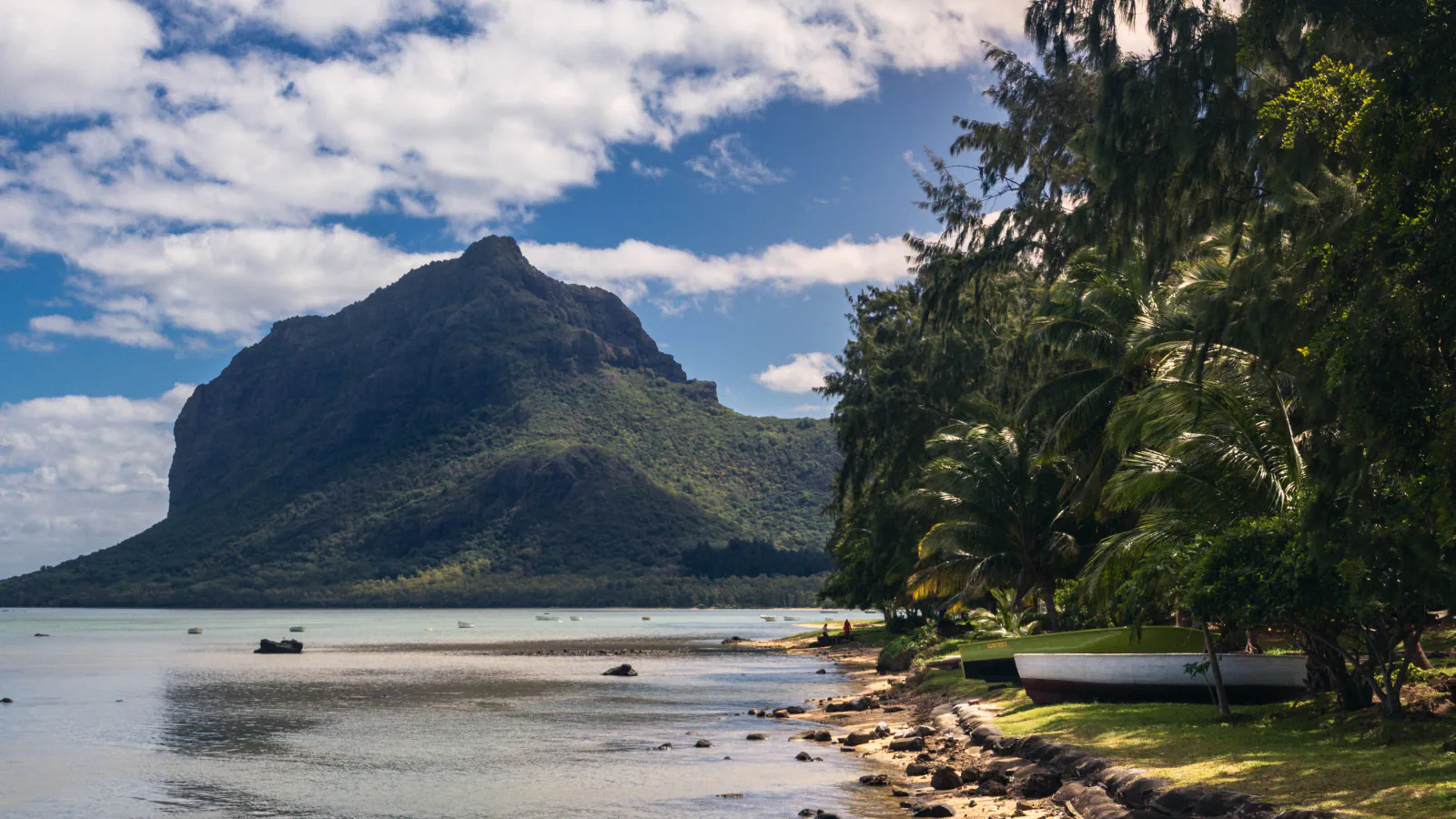 Le Morne Brabant rises beyond a shallow lagoon, with boats moored along the palm-lined shore.