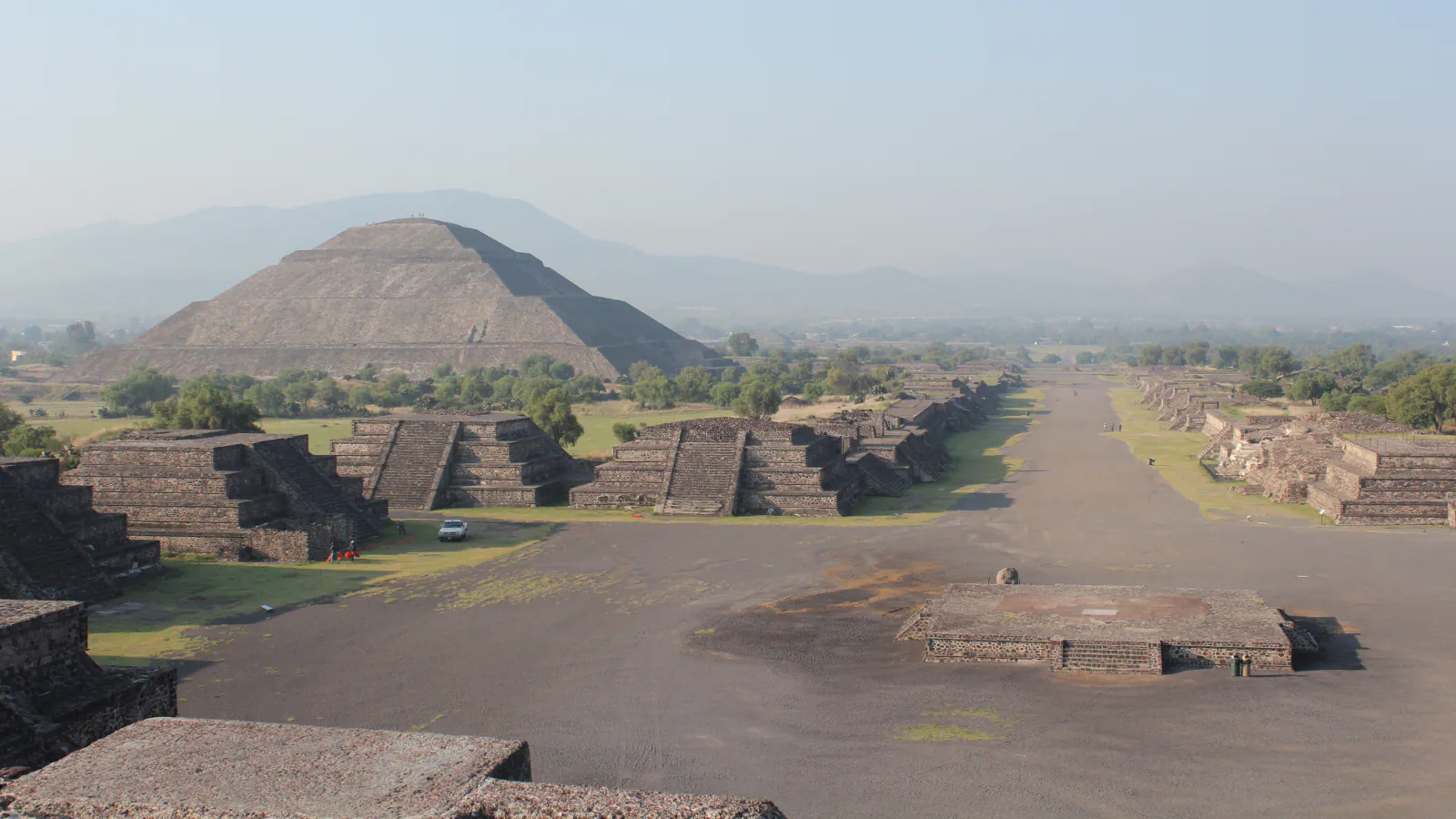 Wide Avenue of the Dead at Teotihuacan on a hazy day, with the Pyramid of the Sun at left and low stepped platforms along the avenue