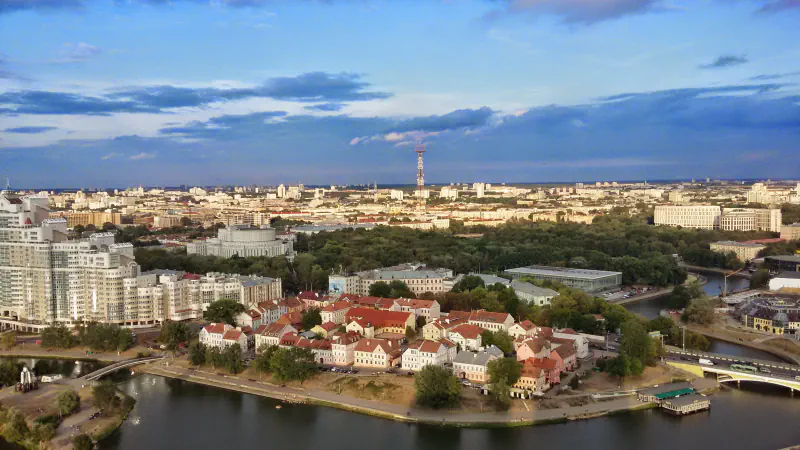 Aerial view of the Svisloch curving past a cluster of red-roofed buildings, with tree cover and city blocks beyond under a cloudy sky