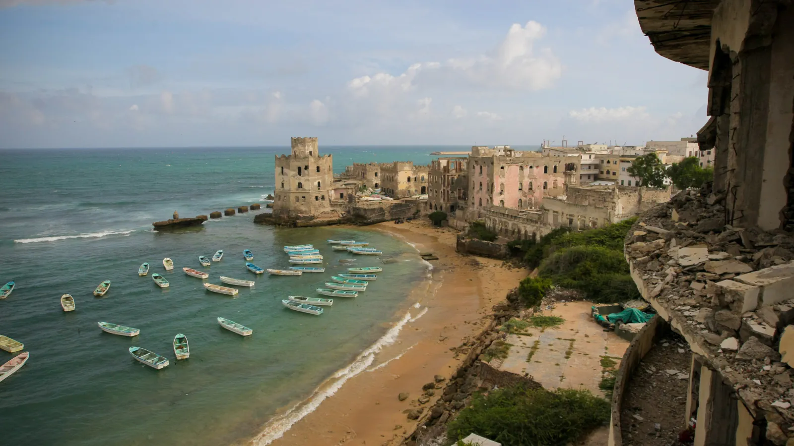 Small fishing boats in a bay beside a sandy beach, with weathered buildings along the shore and a crumbling structure at right