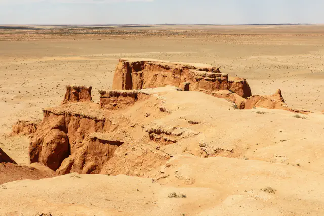Reddish-orange sandstone cliffs and gullies rising from a flat, pale desert plain that reaches the horizon under a hazy sky