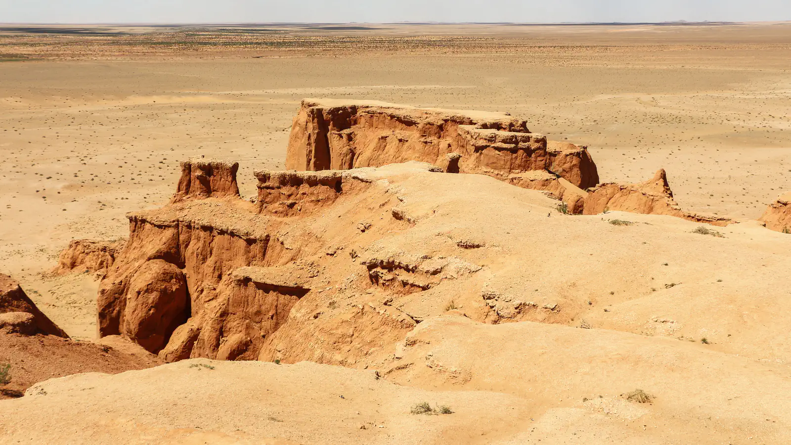Reddish-orange sandstone cliffs and gullies rising from a flat, pale desert plain that reaches the horizon under a hazy sky