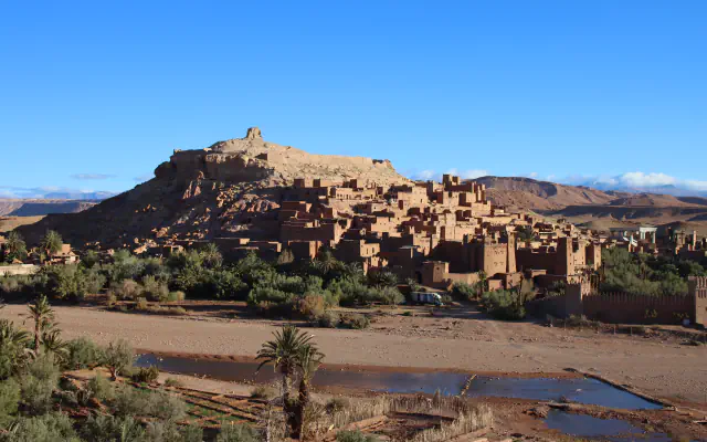 Earthen houses and towers of the ksar of Aït Benhaddou climbing a hillside above a dry riverbed with palms and a shallow pool