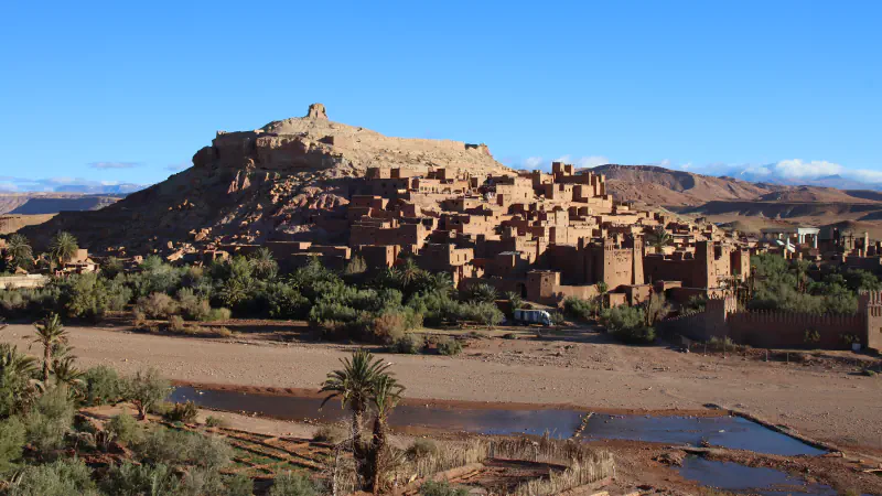 Earthen houses and towers of the ksar of Aït Benhaddou climbing a hillside above a dry riverbed with palms and a shallow pool
