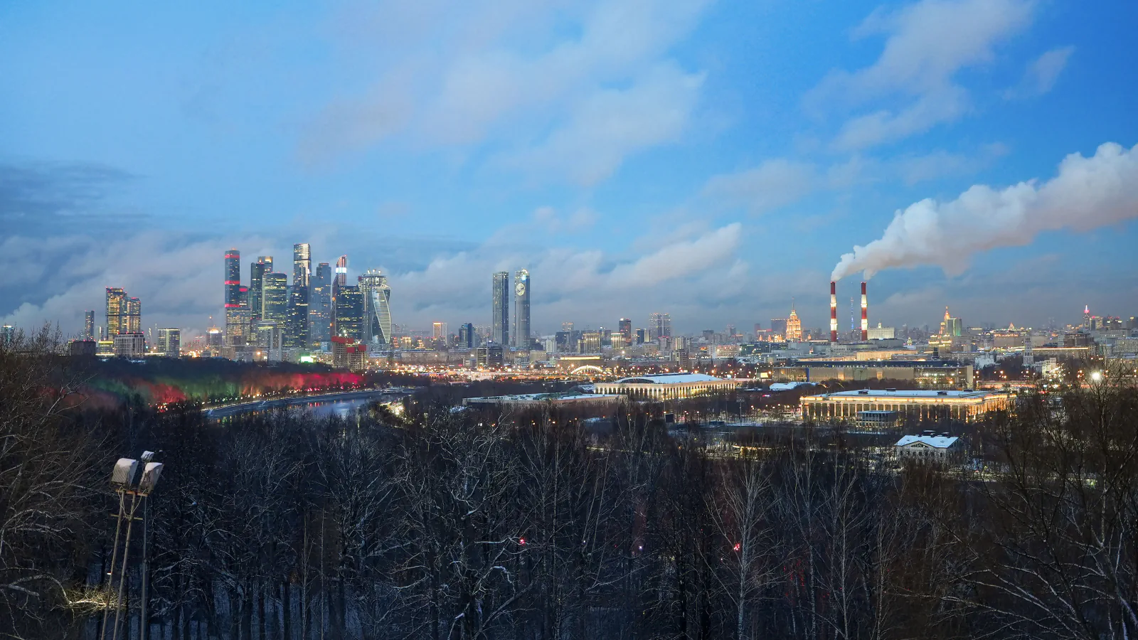 Winter view of Moscow’s skyline at dusk, with towers, a river bend and steam plumes beyond bare trees.