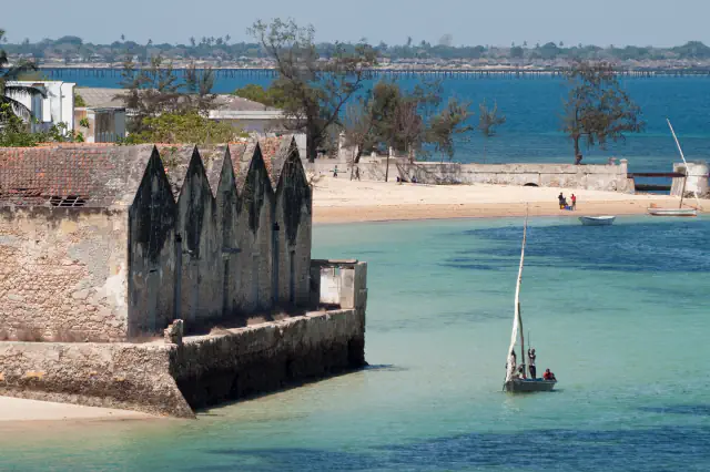 A weathered stone building with a row of pointed gables and a worn tiled roof stands at the water’s edge; a small sailing boat with three people lies on turquoise water, with a sandy beach, a sea wall and a long bridge in the background