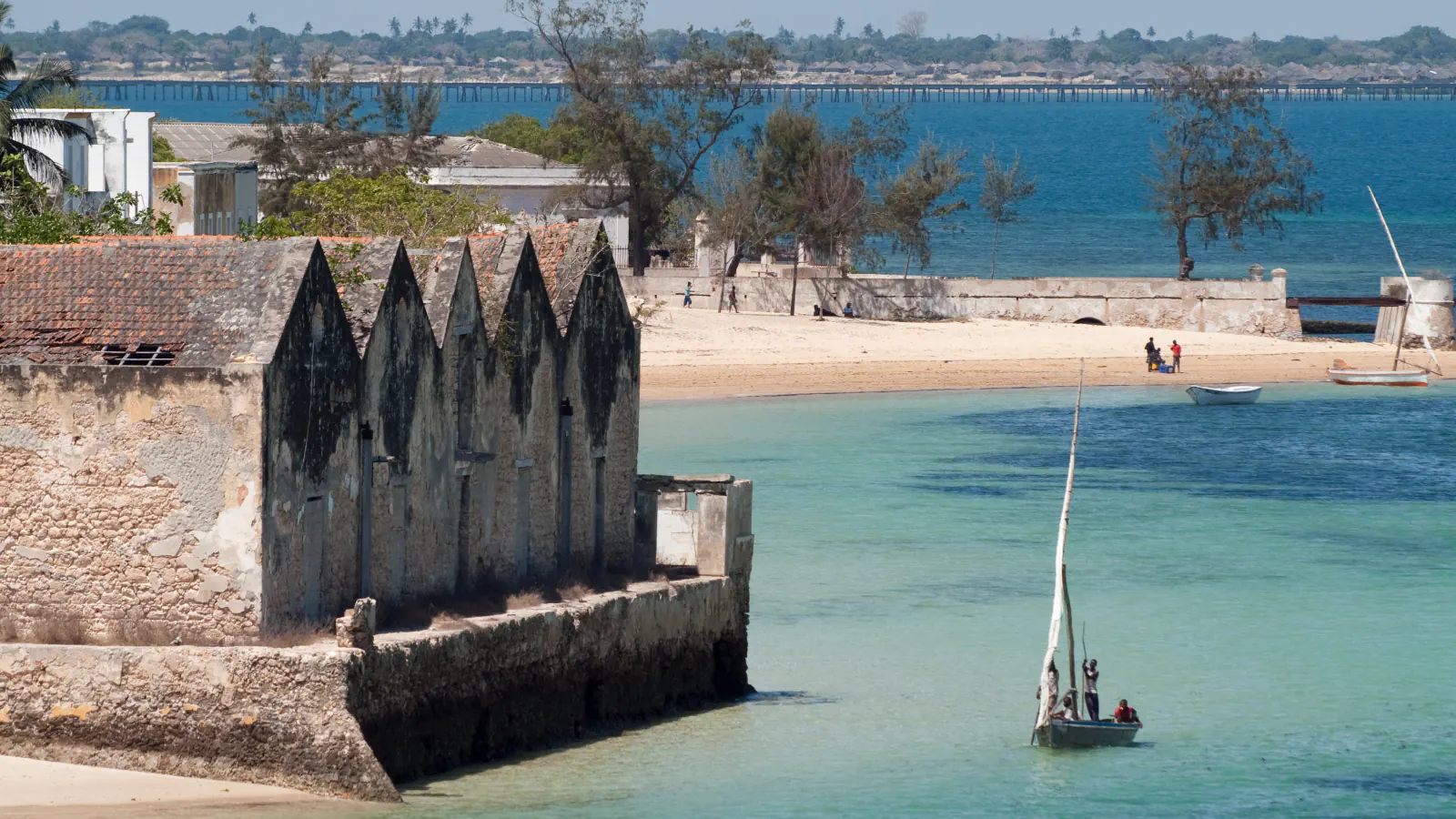 A weathered stone building with a row of pointed gables and a worn tiled roof stands at the water’s edge; a small sailing boat with three people lies on turquoise water, with a sandy beach, a sea wall and a long bridge in the background