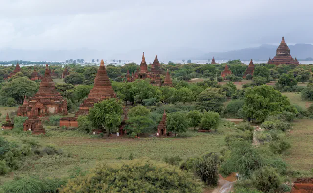 Rdečerjavi opečni templji in stupe se dvigajo nad zelenimi drevesi na ravnini Bagan, v ozadju pa so griči in sivo, oblačno nebo