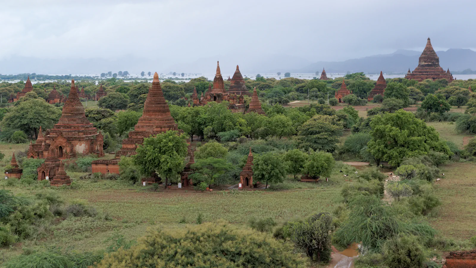 Red-brown brick temples and stupas rising above green trees on the Bagan plain, with hills and a grey, cloudy sky in the distance