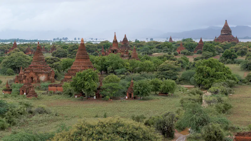 Red-brown brick temples and stupas rising above green trees on the Bagan plain, with hills and a grey, cloudy sky in the distance