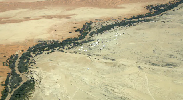 Aerial view of Gobabeb’s white buildings on a pale gravel plain beside the dark green Kuiseb River woodland, with orange dunes beyond