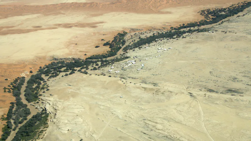 Aerial view of Gobabeb’s white buildings on a pale gravel plain beside the dark green Kuiseb River woodland, with orange dunes beyond