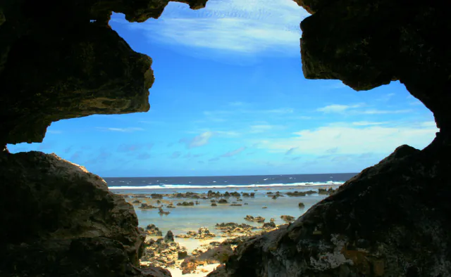 A jagged dark rock opening frames bright blue sky, a line of white surf and a shallow reef flat scattered with low rocks and pale sand
