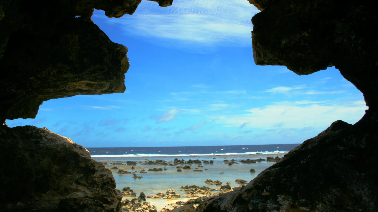 A jagged dark rock opening frames bright blue sky, a line of white surf and a shallow reef flat scattered with low rocks and pale sand