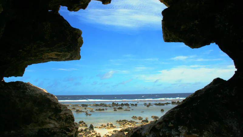 A jagged dark rock opening frames bright blue sky, a line of white surf and a shallow reef flat scattered with low rocks and pale sand