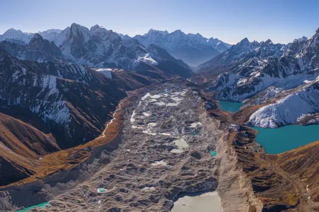 High view down a glacier-filled valley: grey debris-covered ice and pale ponds between brown slopes, turquoise Gokyo lakes on the right and snow-covered peaks beyond