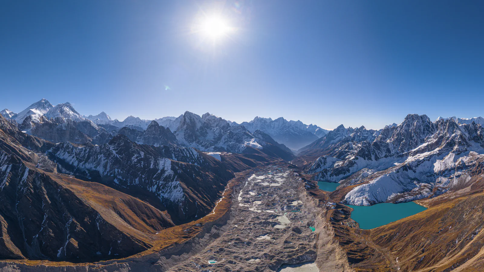 High view down a glacier-filled valley: grey debris-covered ice and pale ponds between brown slopes, turquoise Gokyo lakes on the right and snow-covered peaks beyond