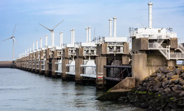 Concrete piers and gate structures of the Eastern Scheldt Barrier along the shore, with wind turbines behind and rock armour on the right