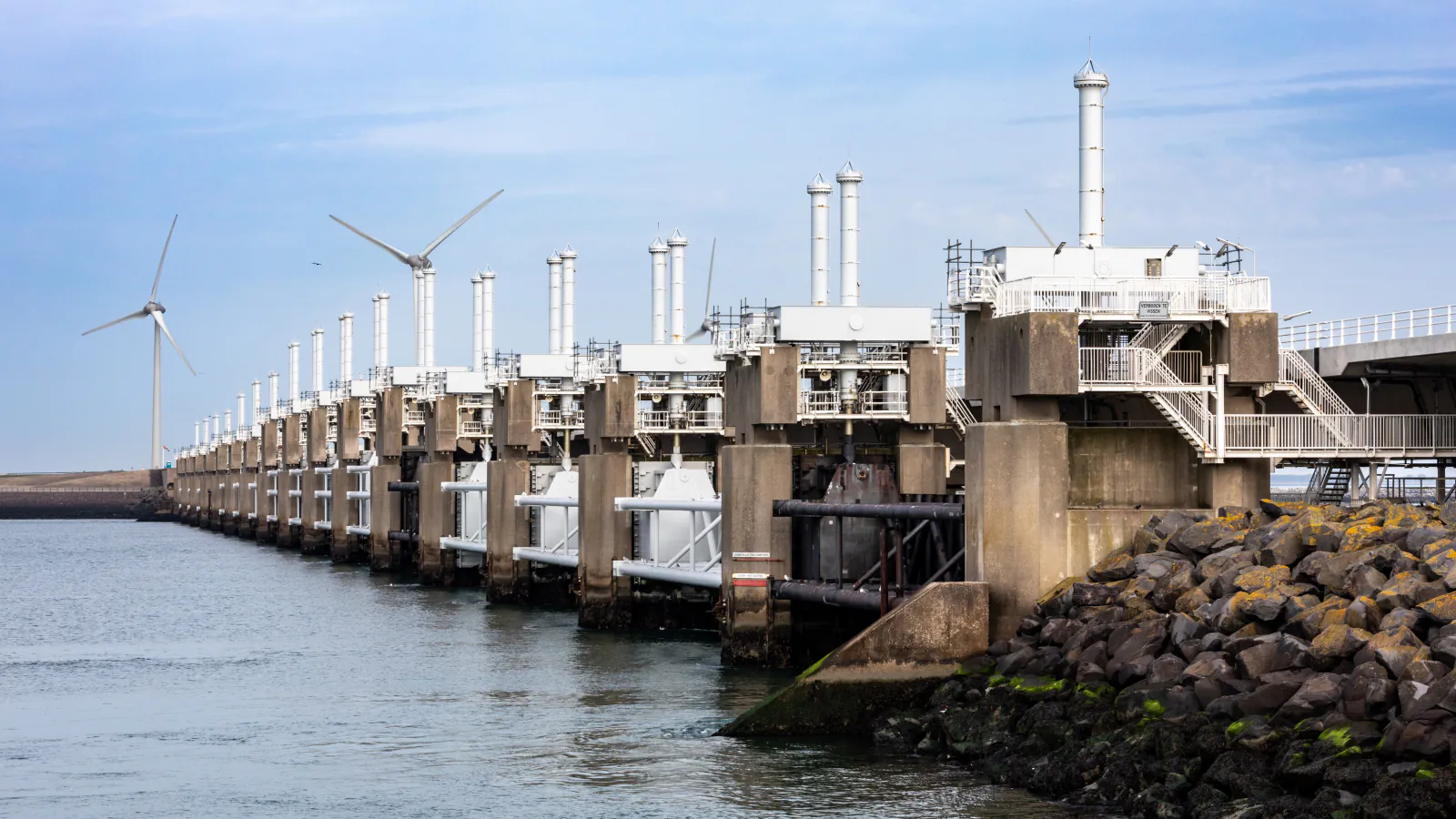 Concrete piers and gate structures of the Eastern Scheldt Barrier along the shore, with wind turbines behind and rock armour on the right