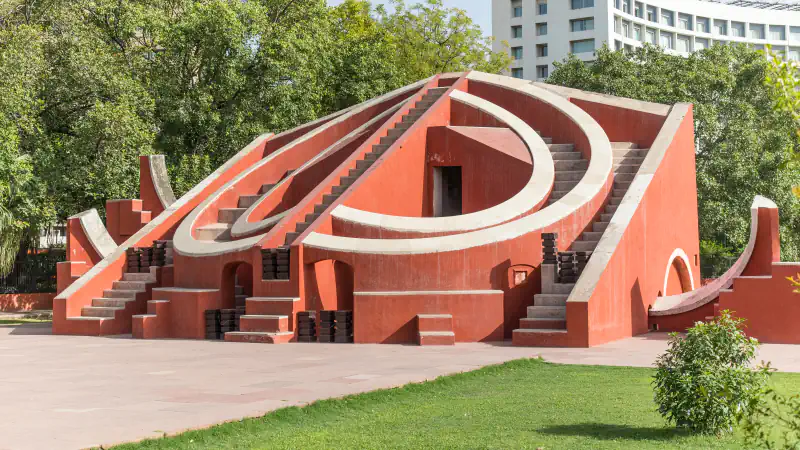 Red curved stepped structure with pale edges at Jantar Mantar, surrounded by trees and lawn.