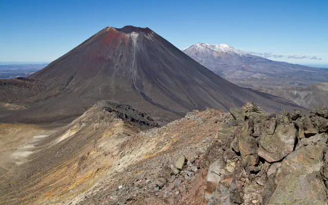 A dark conical volcano with a red-brown crater rim and a snow-dusted mountain beyond it, seen from a rocky ridge in Tongariro National Park