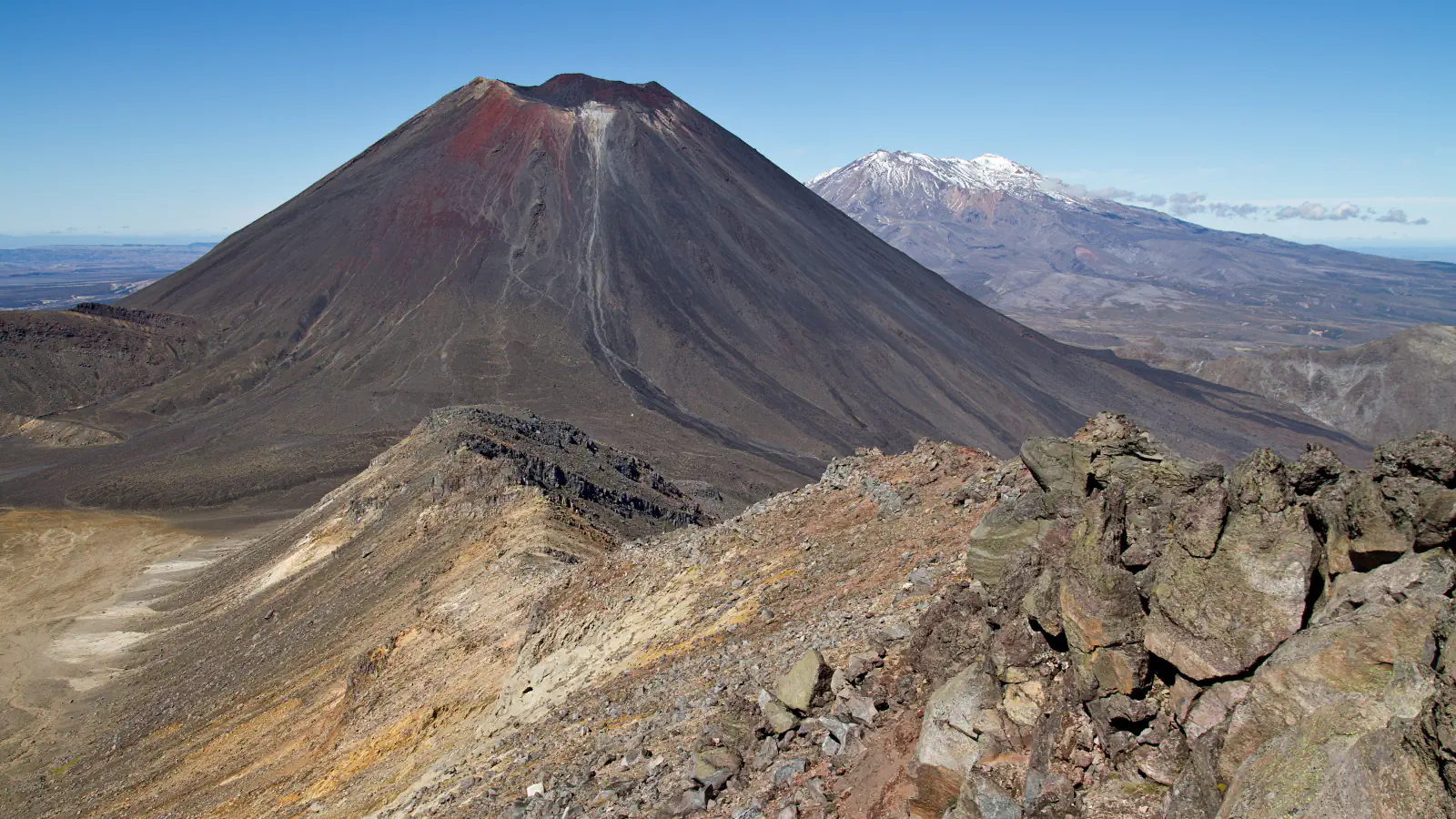 A dark conical volcano with a red-brown crater rim and a snow-dusted mountain beyond it, seen from a rocky ridge in Tongariro National Park