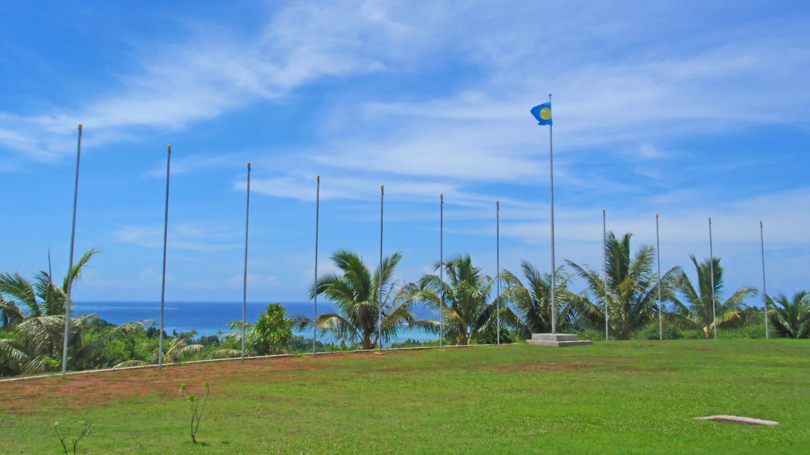 Palau’s flag on one of a row of flagpoles above a lawn, with palms and the sea behind