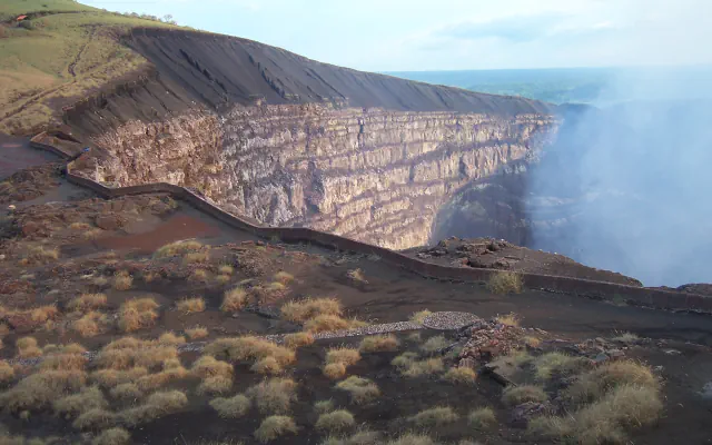 A deep volcanic crater with a layered rock wall, a low stone wall winding along the rim and a pale haze drifting on the right, with tufts of dry grass in the foreground