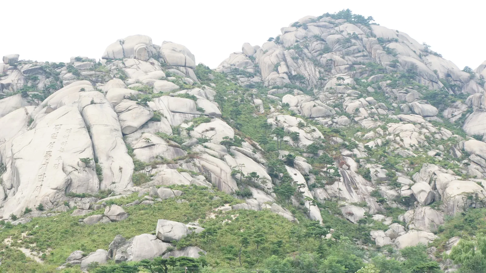 Pale rounded granite slopes dotted with pines under a hazy sky, with Korean characters carved into one rock face