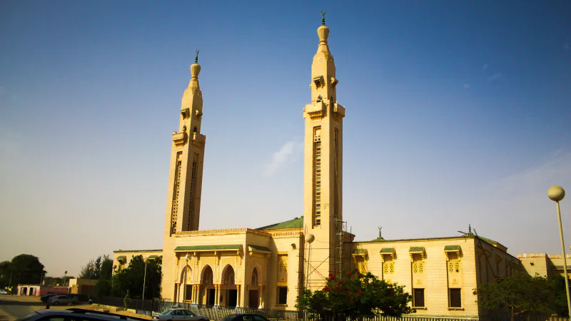 Dva visoka peščeno obarvana minareta nad mošejo z zelenimi strešnimi napušči pod jasnim modrim nebom