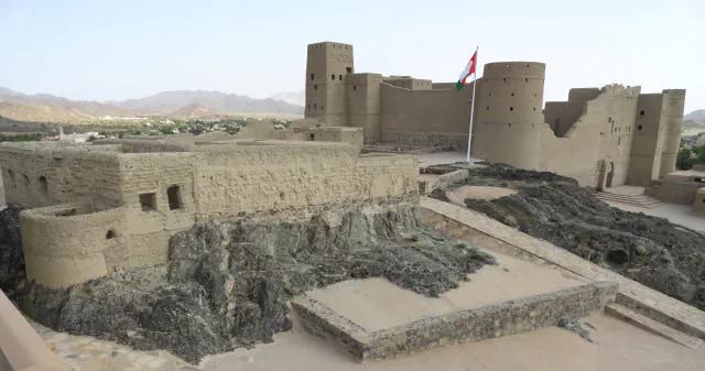 Bahla Fort’s tan walls, a square tower and round towers above dark rock, with an Omani flag and distant hills in view.