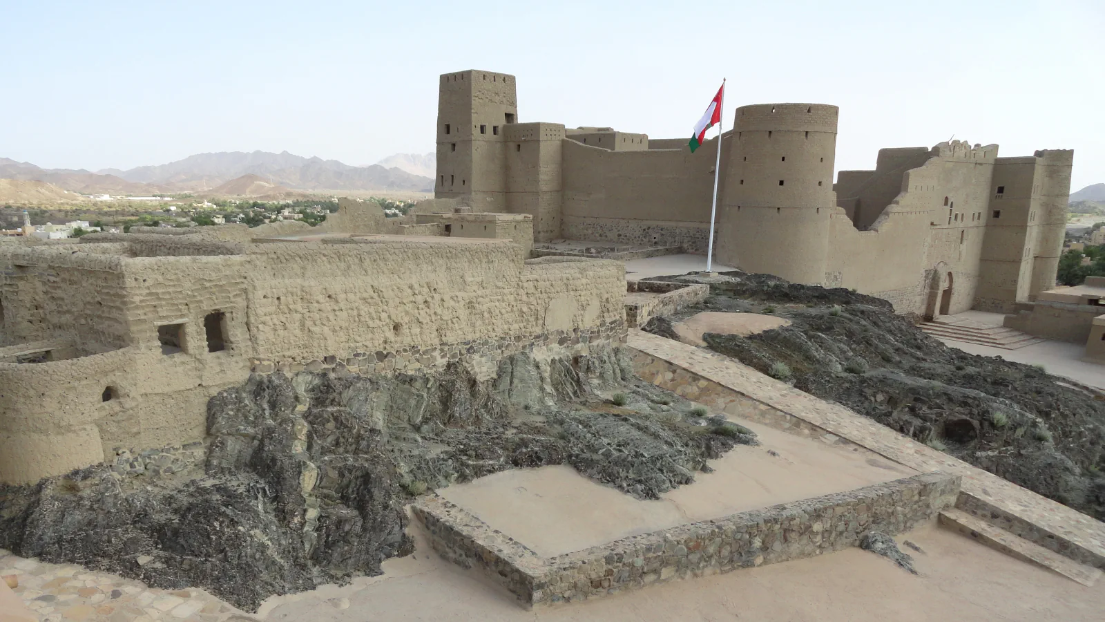 Bahla Fort’s tan walls, a square tower and round towers above dark rock, with an Omani flag and distant hills in view.