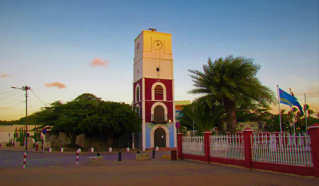 The white, red and blue Willem III Tower in Oranjestad at dusk, with a clock face, trees, a white fence and a flag