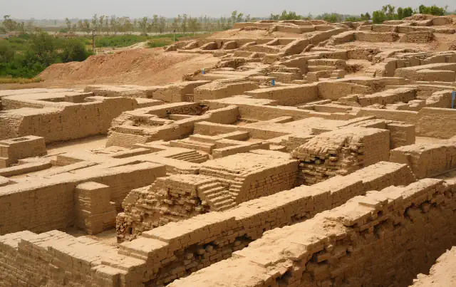 Brick walls, narrow rooms and steps of the excavated city of Mohenjo-daro seen from above, with a mound of earth at left and green fields in the distance