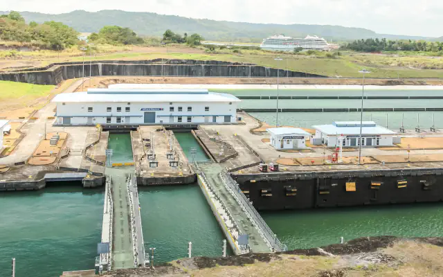 Lock chambers with gate recesses and white service buildings at the Agua Clara Locks, with green hills and a cruise ship in the distance