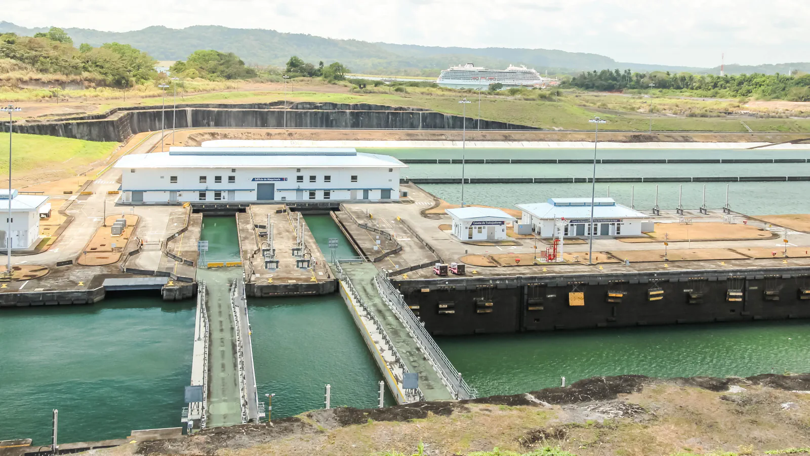 Lock chambers with gate recesses and white service buildings at the Agua Clara Locks, with green hills and a cruise ship in the distance